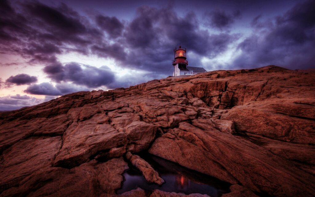 Solitary lighthouse on rugged rocks under violet and amber sky dramatic nature wallpaper background