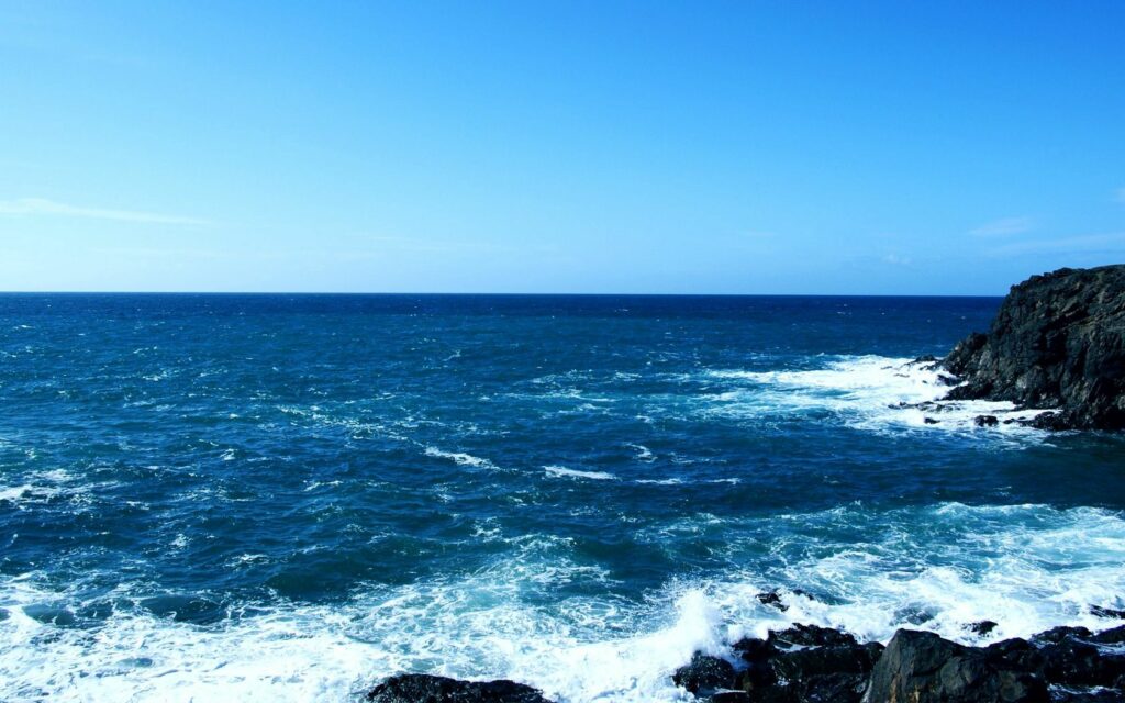Vivid blue ocean waves crashing on dark rugged rocks under clear sky serene sea wallpaper background