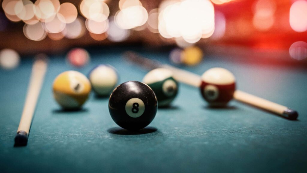 Close-up of black eight-ball on green billiard table with pool cues and vibrant warm bokeh background Wallpaper