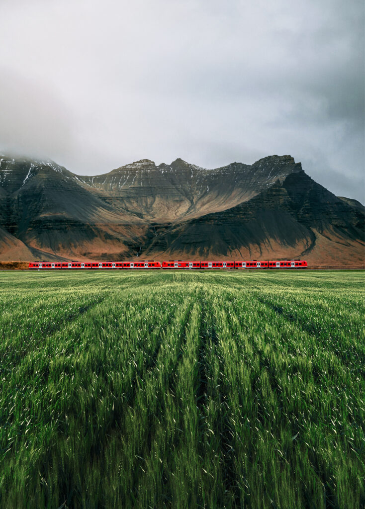 Bright red train crossing lush green field with dark mountains and cloudy sky scenic wallpaper
