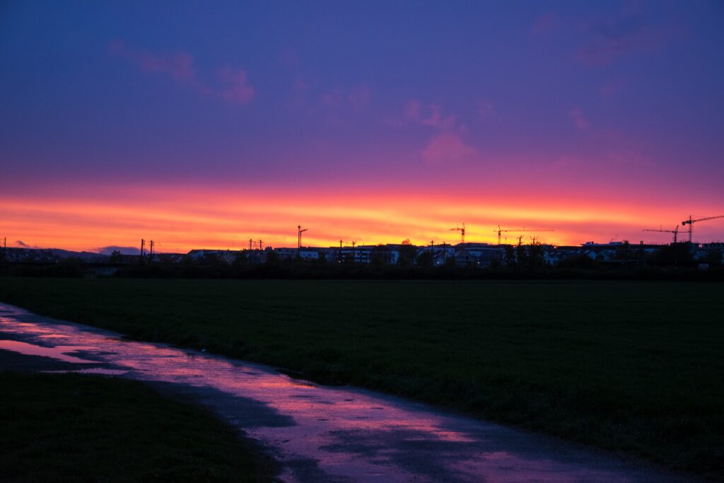 Vivid sunset with orange pink sky over silhouetted buildings cranes trees and reflective winding path Wallpaper