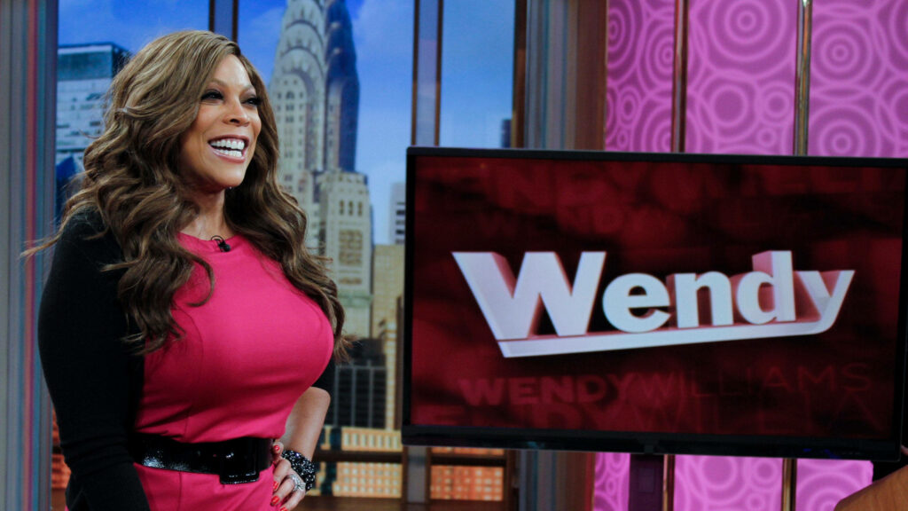 Wendy Williams smiling on talk show set beside Wendy logo screen with vibrant cityscape background Wallpaper