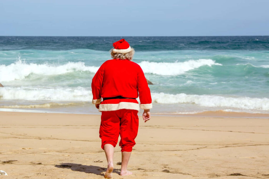 Santa Claus Strolls Barefoot on Sunny Beach | Whimsical Holiday Scene Wallpaper