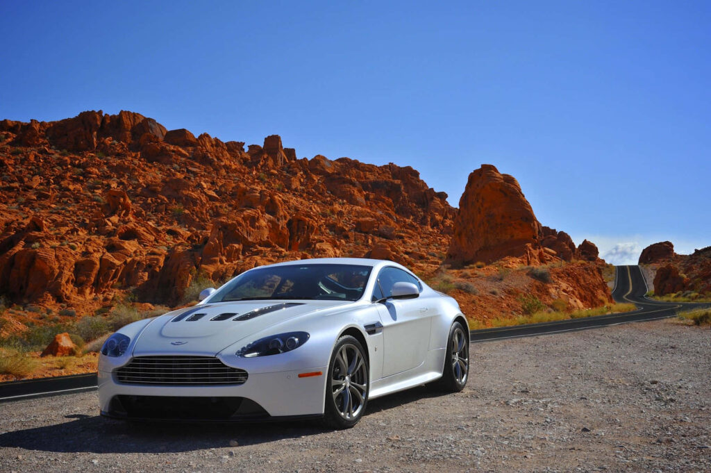 White Aston Martin V12 Vantage on scenic desert road with red rocks and clear blue sky background Wallpaper