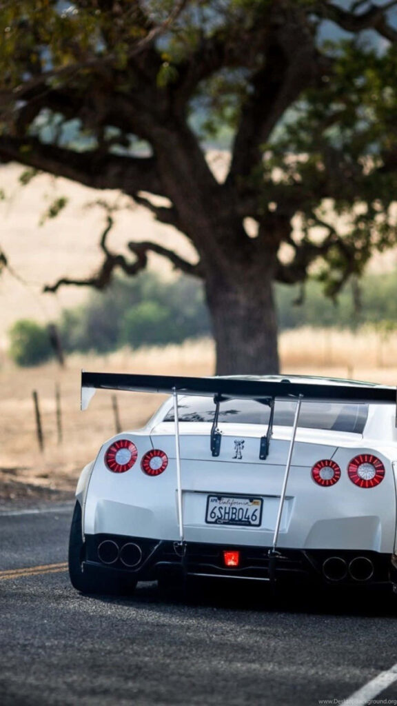 White GTR R35 sports car with large rear wing speeding on winding country road in sunlight Wallpaper