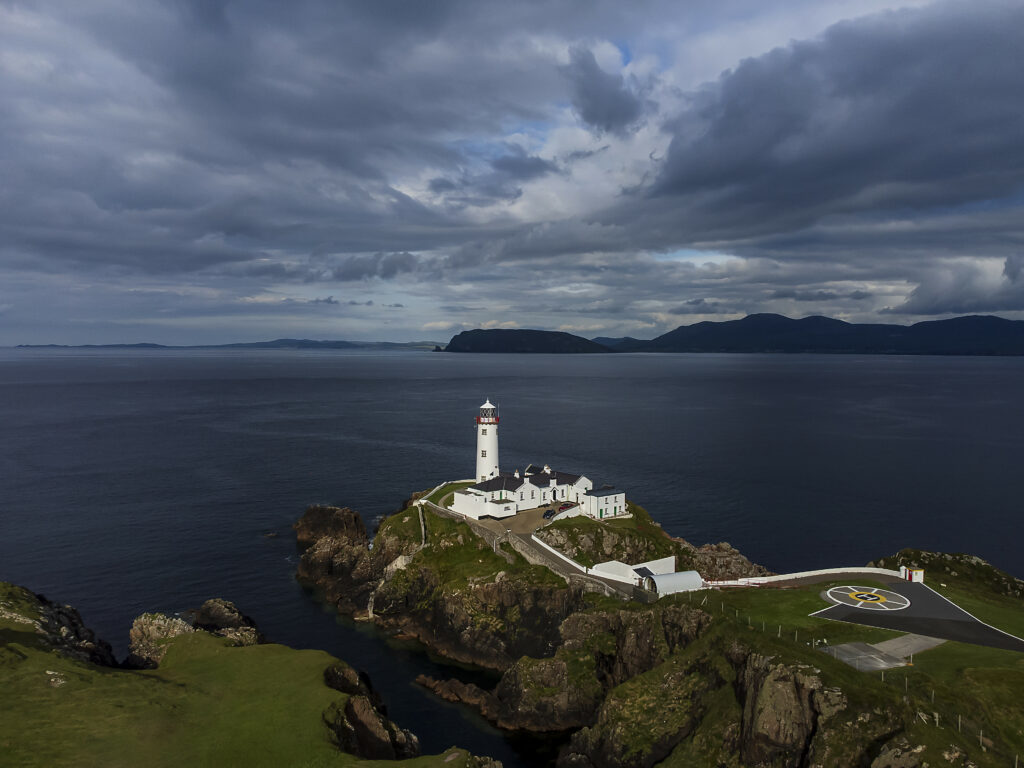 White lighthouse and coastal buildings on rugged cliff with deep blue sea and dramatic sky wallpaper