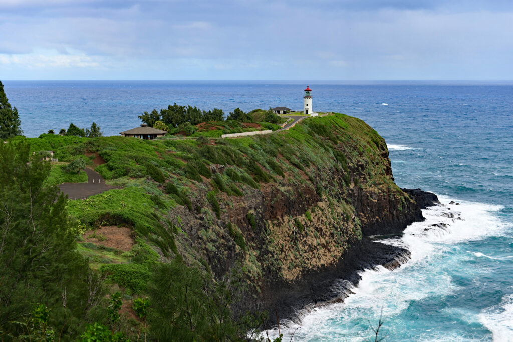 White lighthouse on green cliff overlooking blue sea waves crashing under partly cloudy sky wallpaper