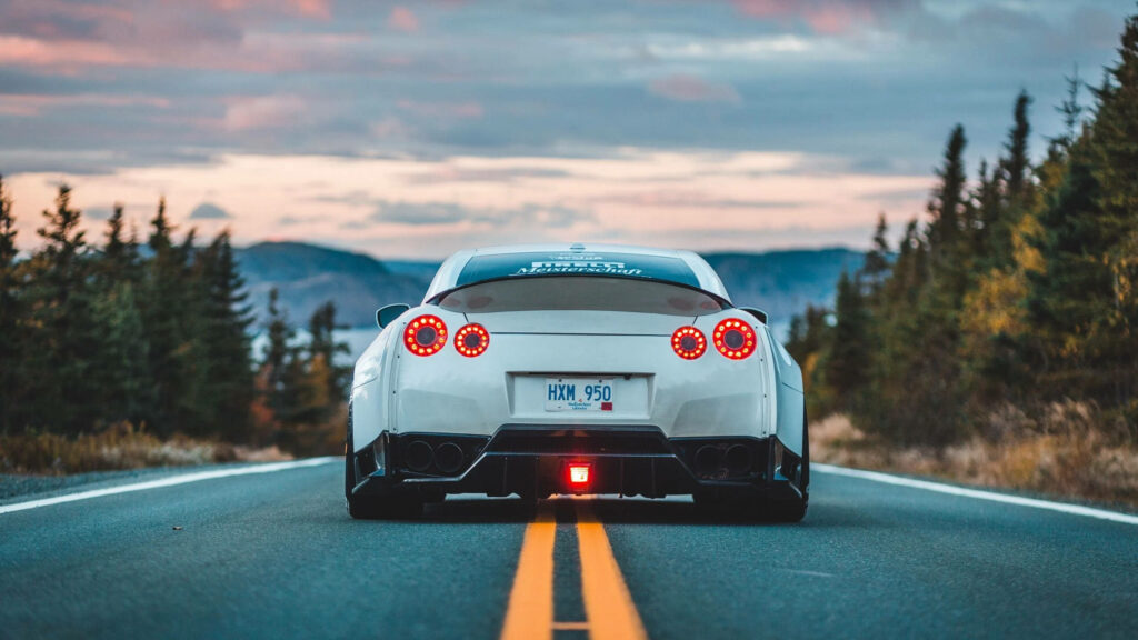 Rear view of white Nissan GT-R on open road with pine trees and colorful sunset sky background Wallpaper