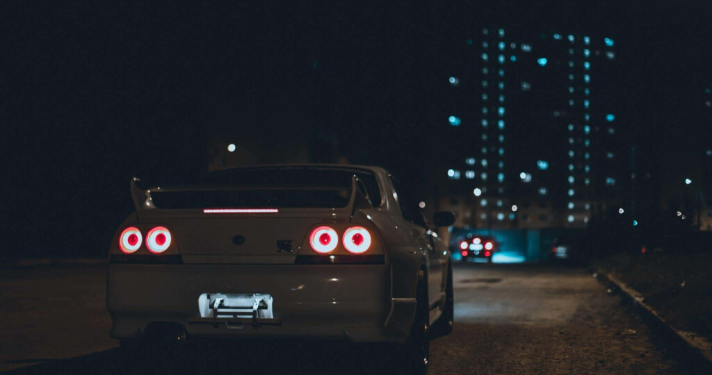 White Nissan R33 GTR parked on dim street with glowing city lights nighttime wallpaper background