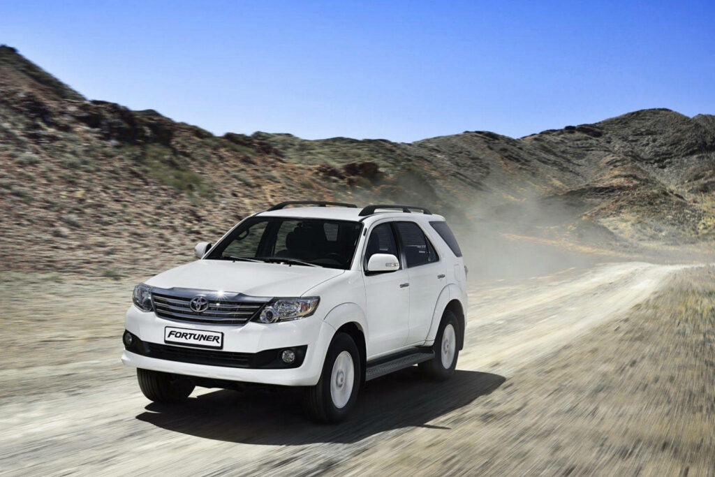 White Toyota Fortuner driving on dusty desert road under clear blue sky rugged landscape wallpaper