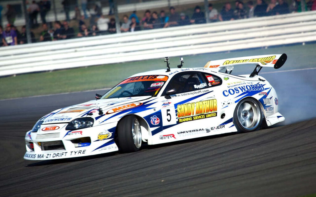 White Toyota Supra race car drifting fast with sponsor decals and tire smoke on race track Wallpaper