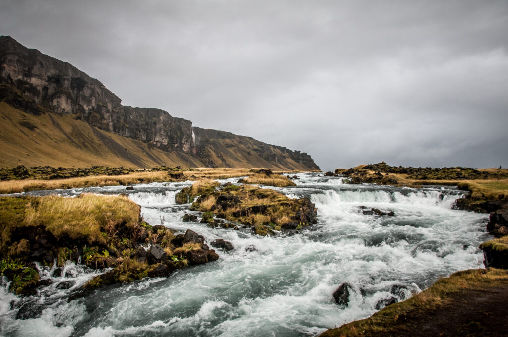Rushing river flows over dark rocks through yellow green grasslands under stormy skies with cliffs Wallpaper