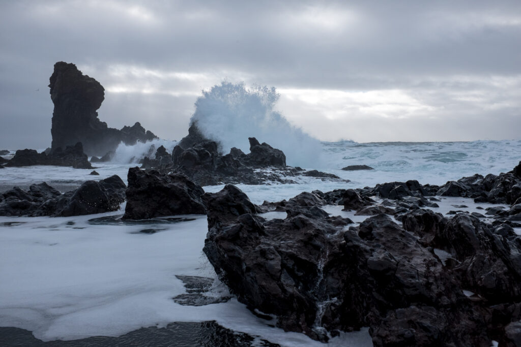 Dark volcanic rocks and crashing sea waves under dramatic cloudy sky on rugged coastline Wallpaper