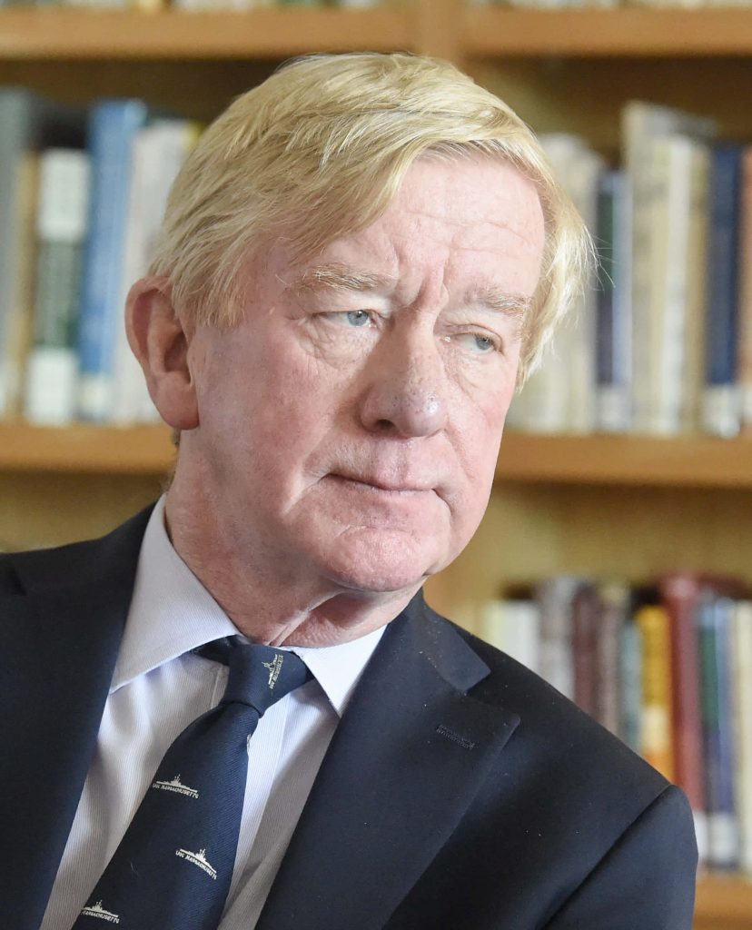 Portrait of William Weld in navy suit by bookshelves with soft natural light and shallow depth Wallpaper