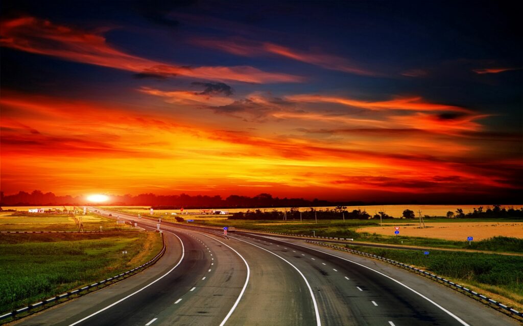Winding asphalt road through green fields at sunset with vivid orange sky and roadside traffic signs Wallpaper