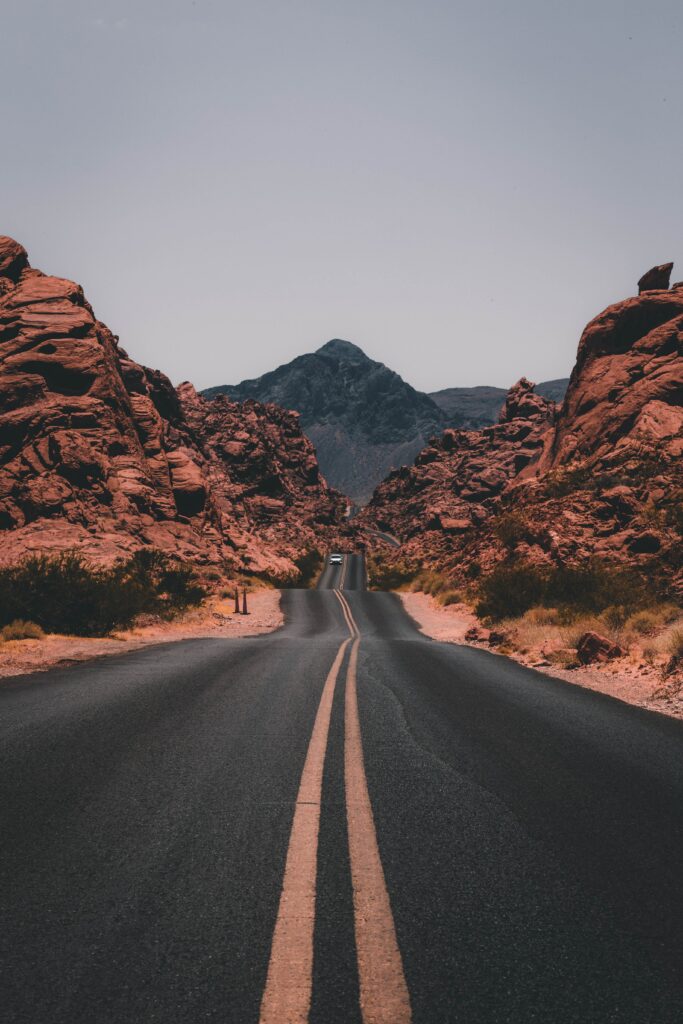 Winding asphalt road with yellow lines through red rock valley toward distant mountain under hazy sky Wallpaper