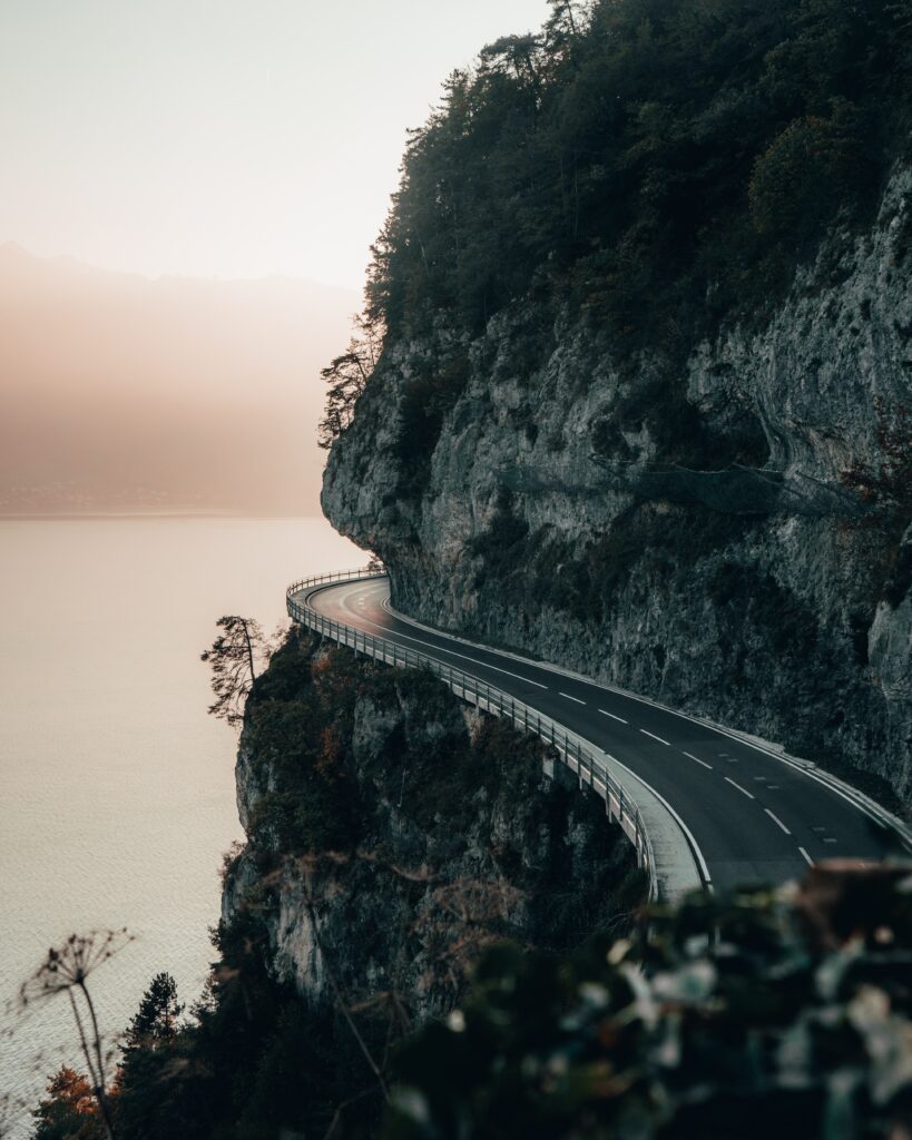Winding cliffside road overlooking calm sea at sunset with pastel sky and rugged rocks Wallpaper