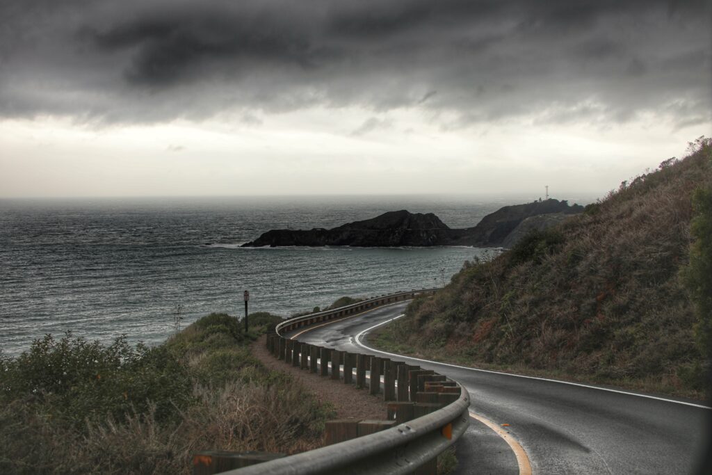 Winding coastal road beside dark ocean with green hills cliffs and dramatic cloudy sky wallpaper