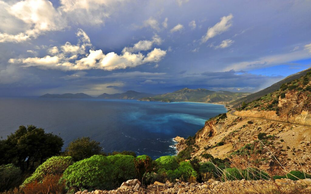 Winding coastal road along rugged cliffs with lush greenery deep blue sea and dramatic sky Wallpaper