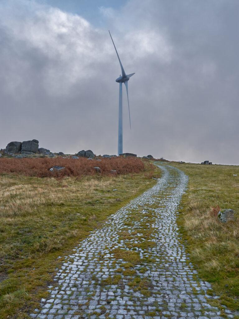 Winding cobblestone path through grassy field to tall windmill with cloudy sky and rocks background Wallpaper