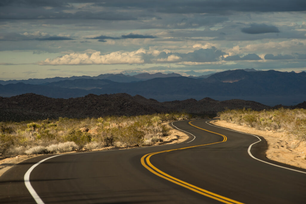 Winding desert road with yellow lines curving through sunlit hills under dramatic sky background Wallpaper
