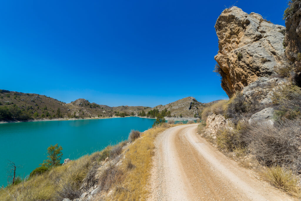 Winding dirt road along turquoise river with rugged mountains rocky cliffs under blue sky wallpaper