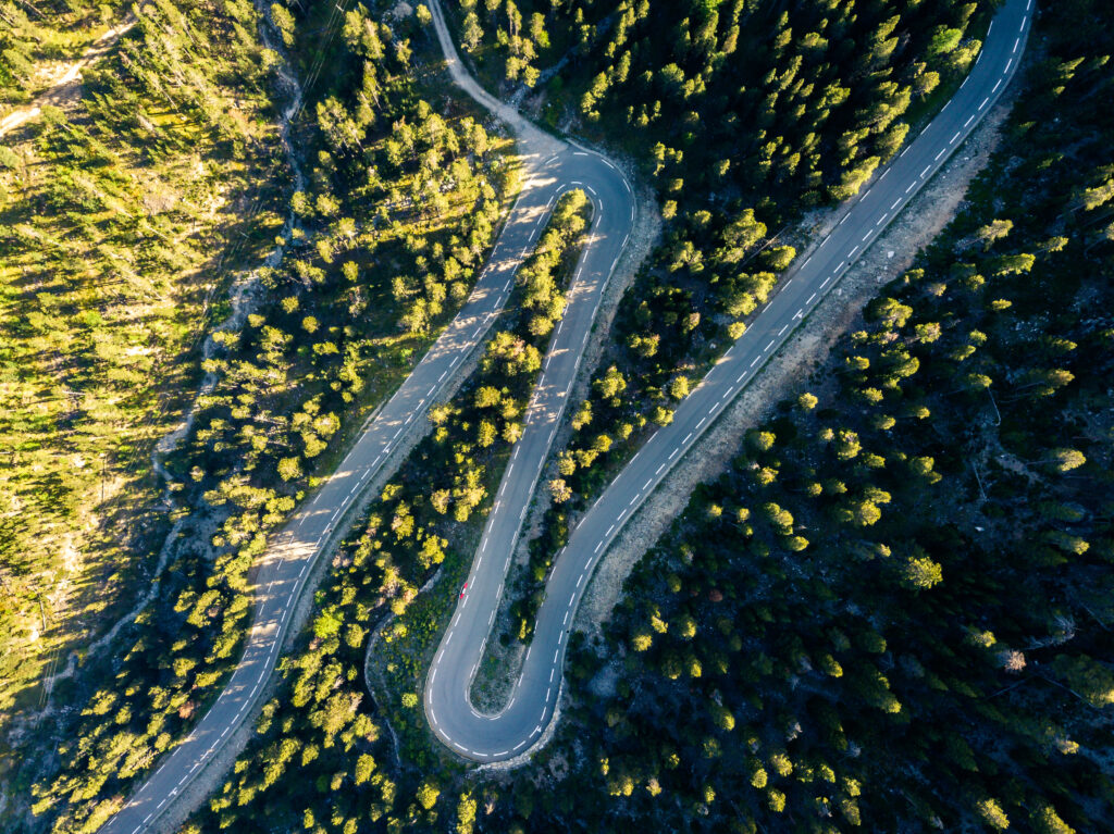 Aerial view of winding road through dense green forest with sunlight casting long shadows wallpaper