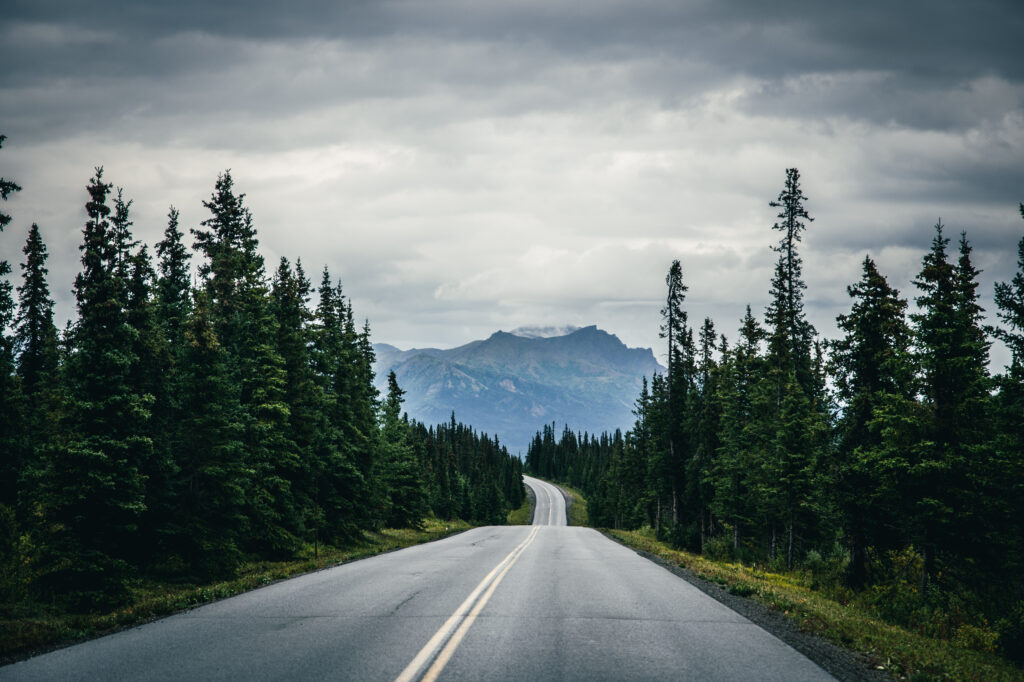 Winding road through dense green forest toward rugged mountains under cloudy sky nature wallpaper