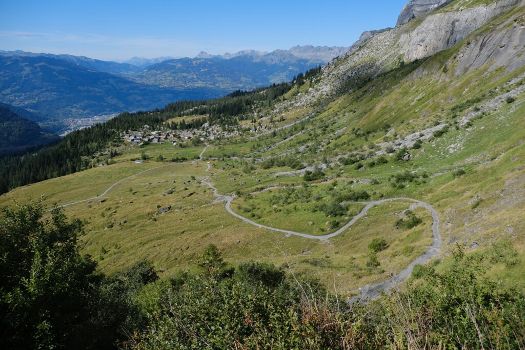 Winding path through lush mountain slope and green valley with houses under vibrant alpine sky Wallpaper