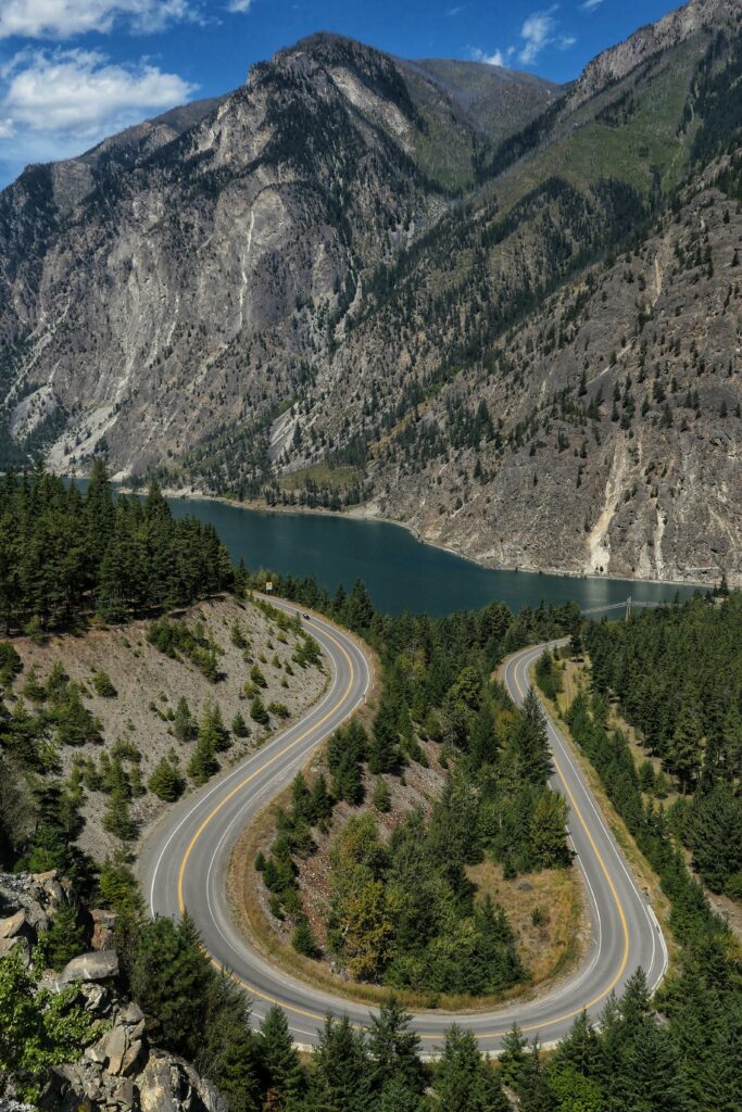 Aerial view of winding road through green forest with mountains and blue lake in background wallpaper