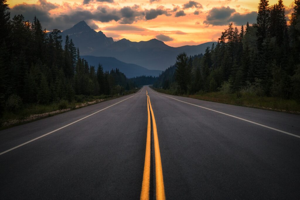 Winding asphalt road with yellow lines through lush trees toward Jasper Canada mountains at sunset Wallpaper