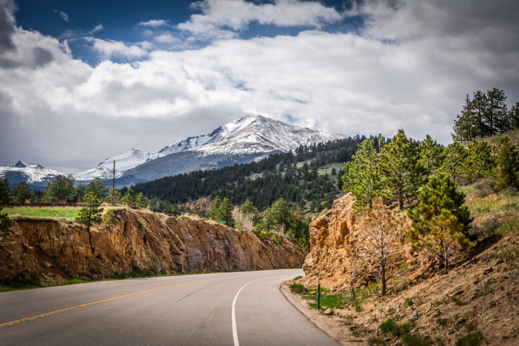 Winding road through rocky cliffs and pine trees leading to snow-capped mountain under cloudy sky Wallpaper