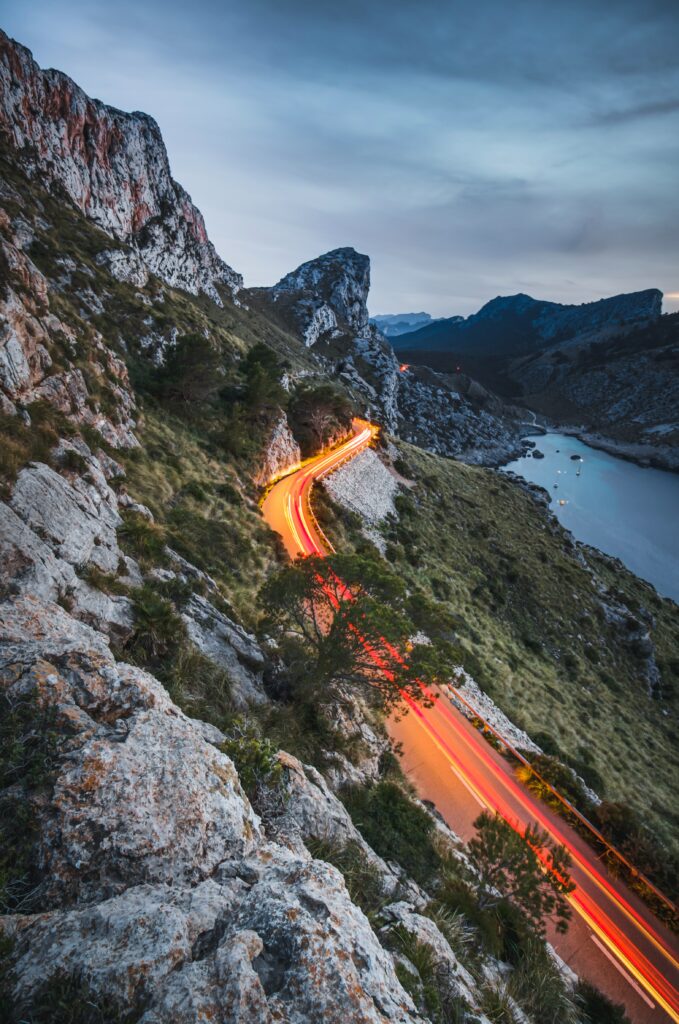 Winding mountain road with vibrant light trails by cliffs and serene lake long exposure landscape Wallpaper