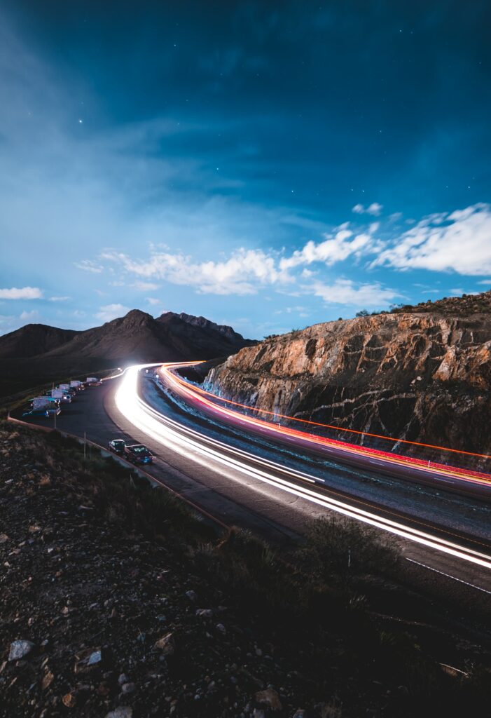 Winding mountain road at night with glowing car light trails and rugged rocky cliffs wallpaper background