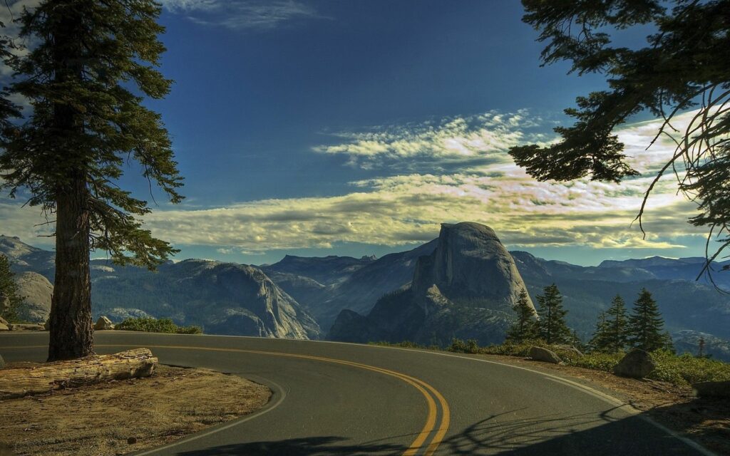 Winding mountain road surrounded by tall trees and rocky cliffs under vibrant blue sky wallpaper