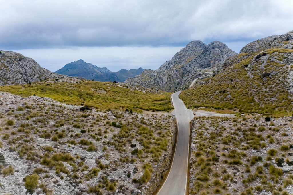 Winding mountain road with rocky hills and grass beneath dramatic cloudy sky serene landscape wallpaper