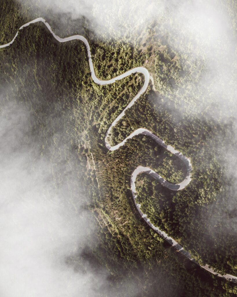 Aerial view of winding mountain road through lush green forest with soft drifting clouds wallpaper