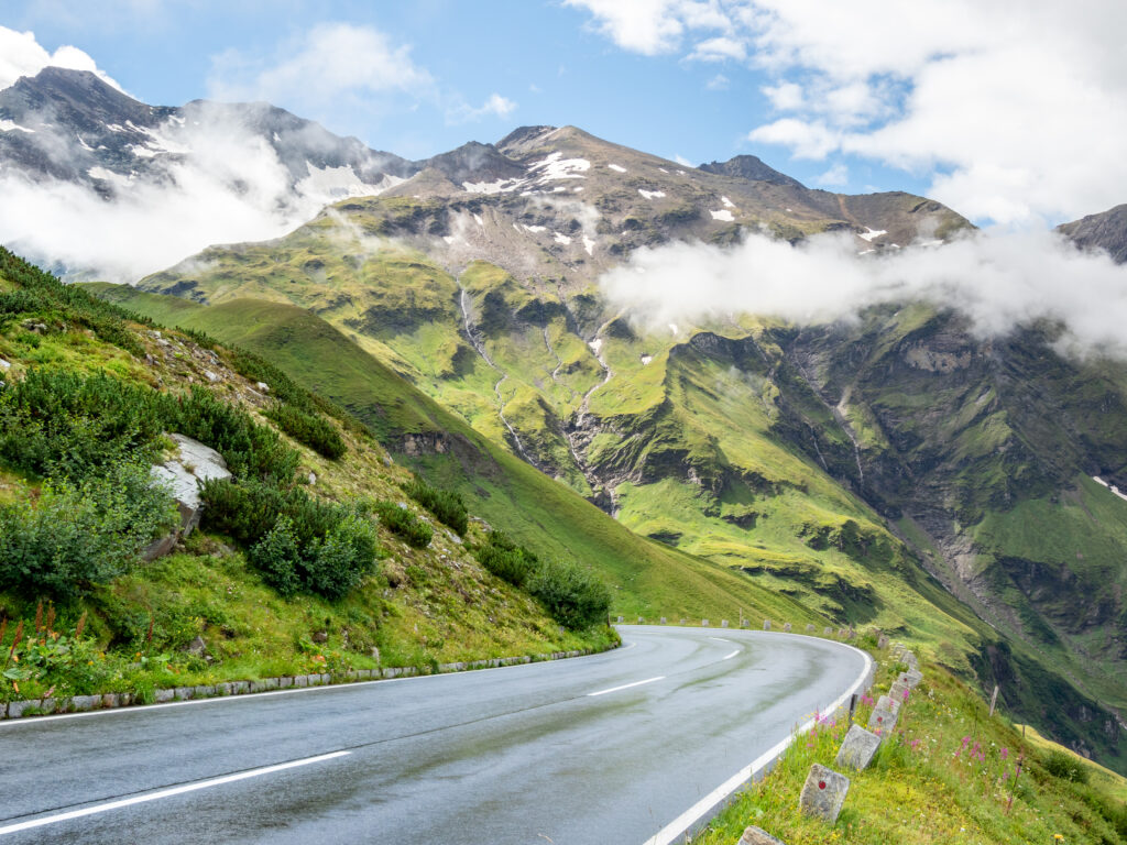 Winding road through lush green grass and rolling mountains under fluffy clouds serene landscape Wallpaper