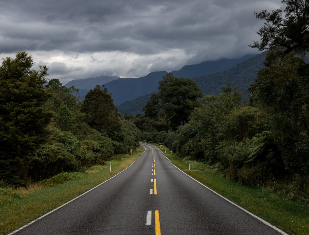 Winding road with yellow markings through green trees under moody sky and distant mountains Wallpaper