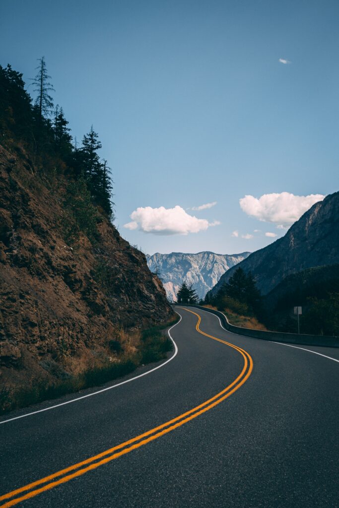Winding asphalt road through rugged mountains with rocky cliffs and scattered trees under blue sky Wallpaper