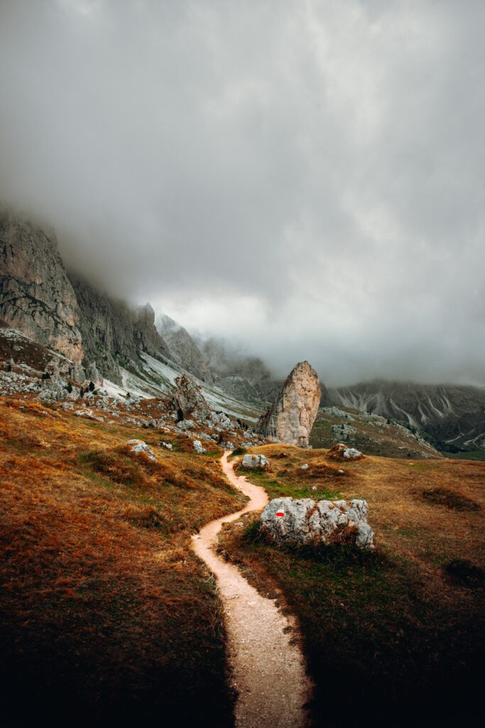 Winding trail through rocky grassy terrain leads to towering mountains beneath dramatic clouds Wallpaper