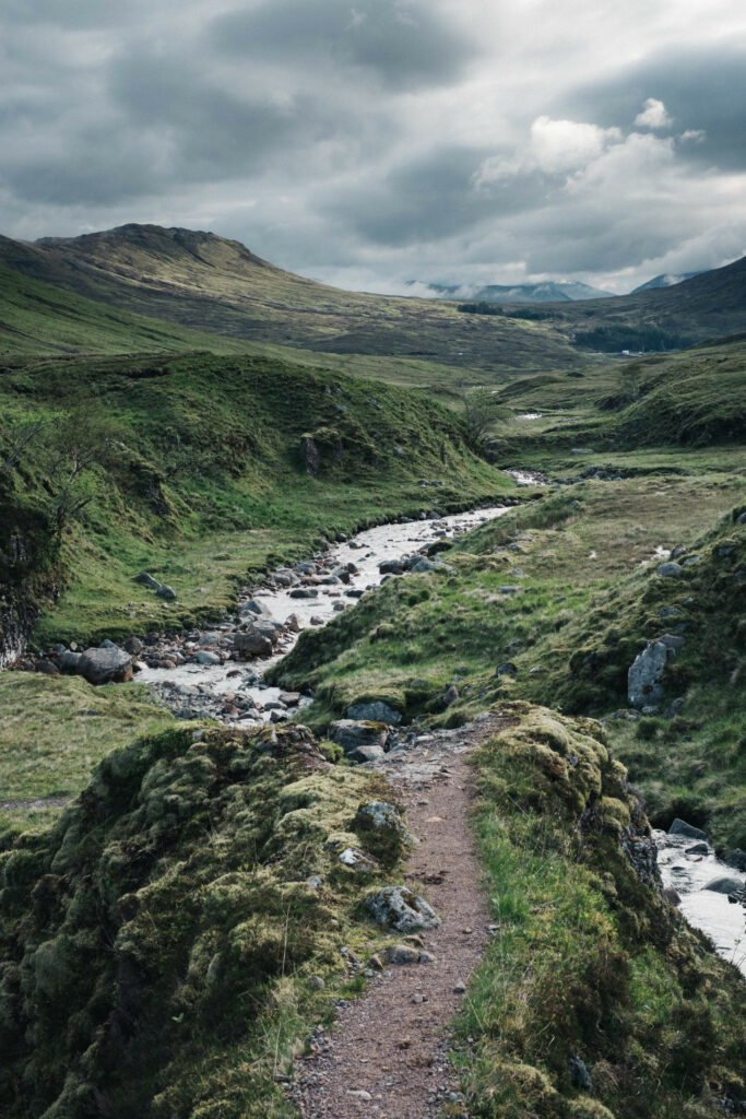 Winding path along gentle stream in lush green valley with mossy stones under cloudy sky Wallpaper