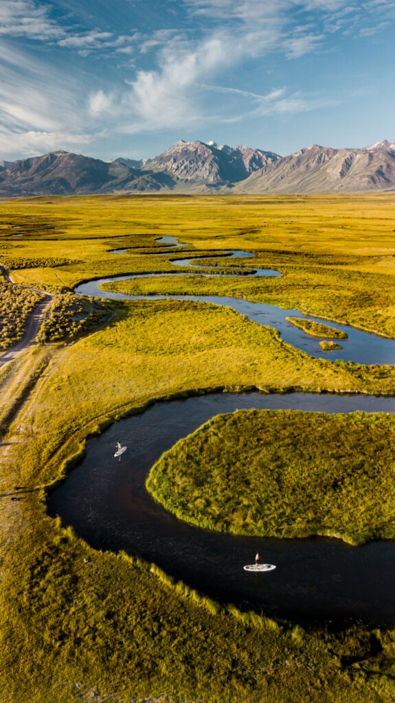Aerial view of winding river through golden fields and mountains with paddleboarders serene nature wallpaper