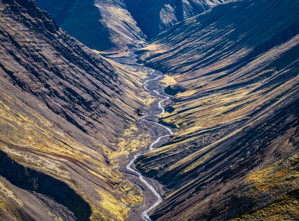 Winding river through rugged mountain valleys with sunlight highlighting rocky slopes and greenery Wallpaper