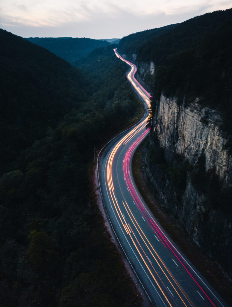 Winding road with car light trails through dark forested valley and high cliffs at night Wallpaper