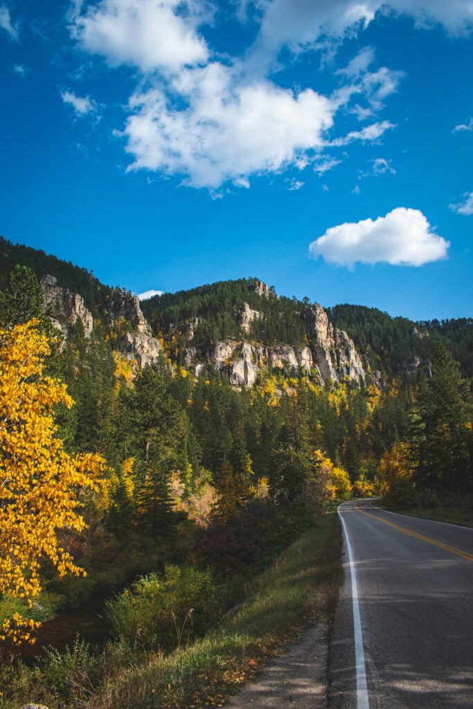Winding road through vibrant autumn trees and jagged rocks with mountains under blue sky wallpaper