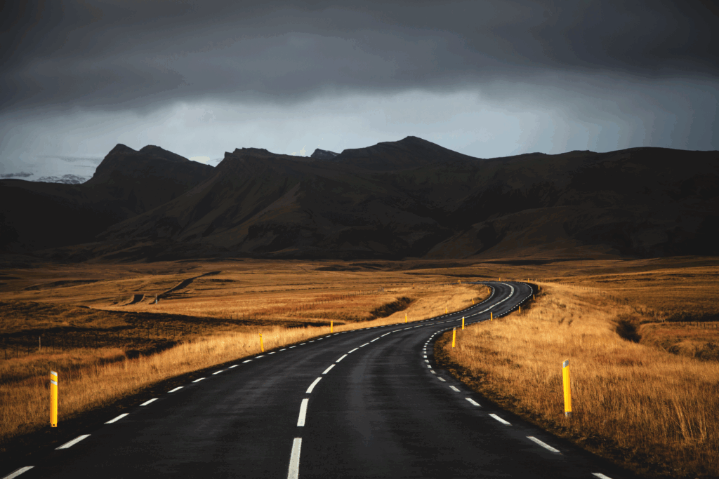 Winding road through golden fields leading to shadowy mountains under moody twilight sky wallpaper