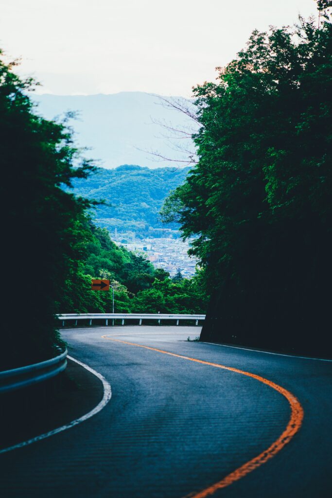 Winding road with sharp turn through lush green trees toward blue mountains under clear sky Wallpaper