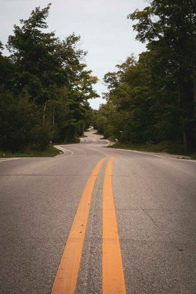 Winding road with bright yellow lines bordered by green trees under overcast sky wallpaper background