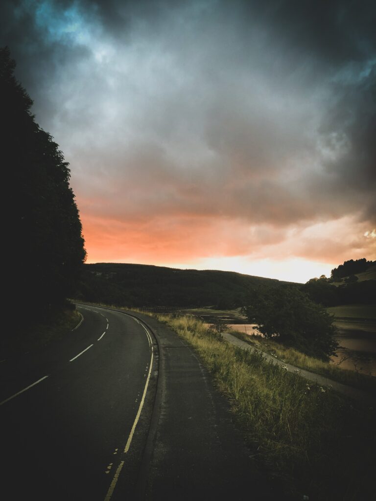 Winding road through lush green trees under dramatic sunset sky with glowing clouds wallpaper
